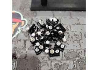 A pile of black fingerboard trucks with white axle nuts on a grey surface.