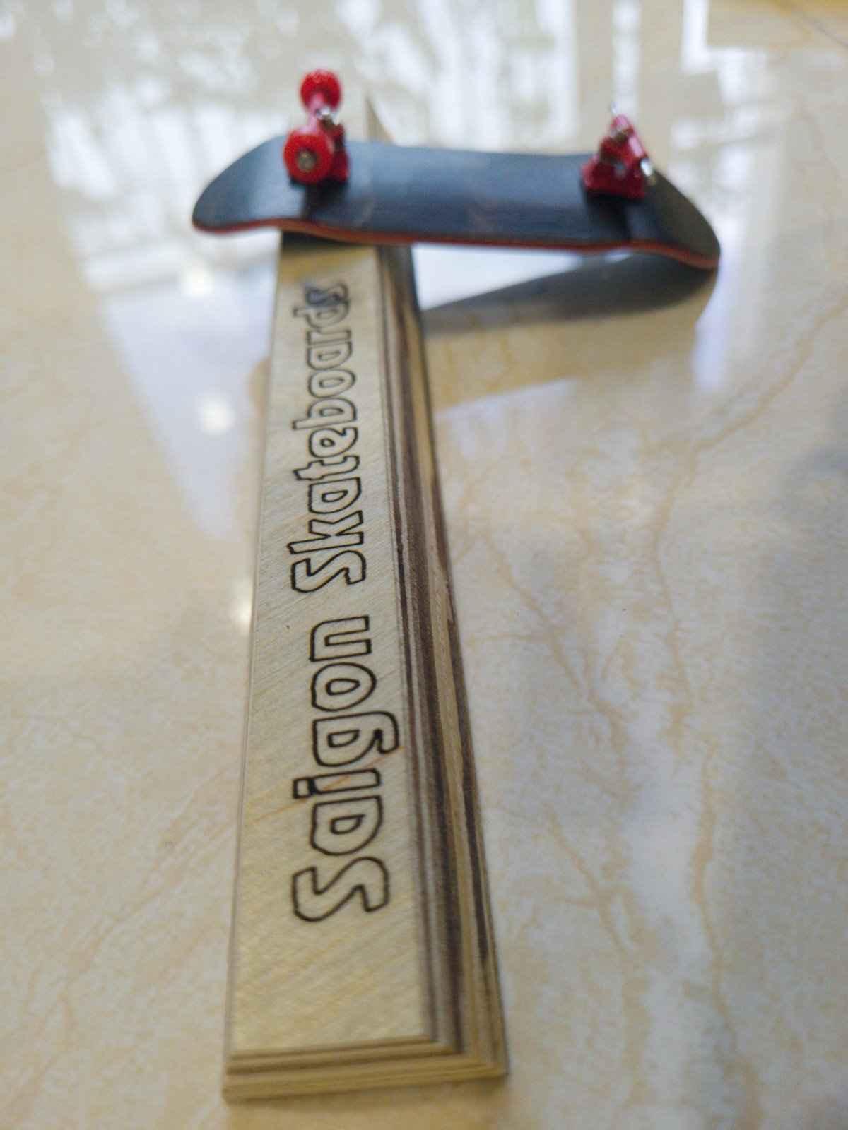 A handmade fingerboard with red wheels resting on a Saigon Skateboards wooden display stand.