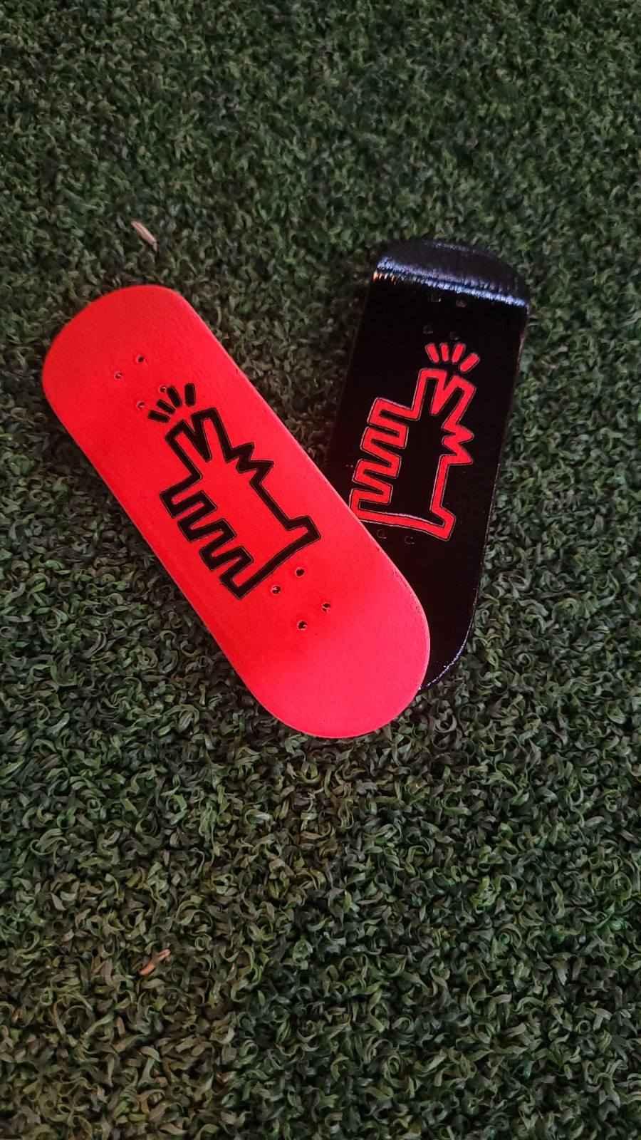 Two custom fingerboard decks, one red and one black, with a graphic design on green turf.