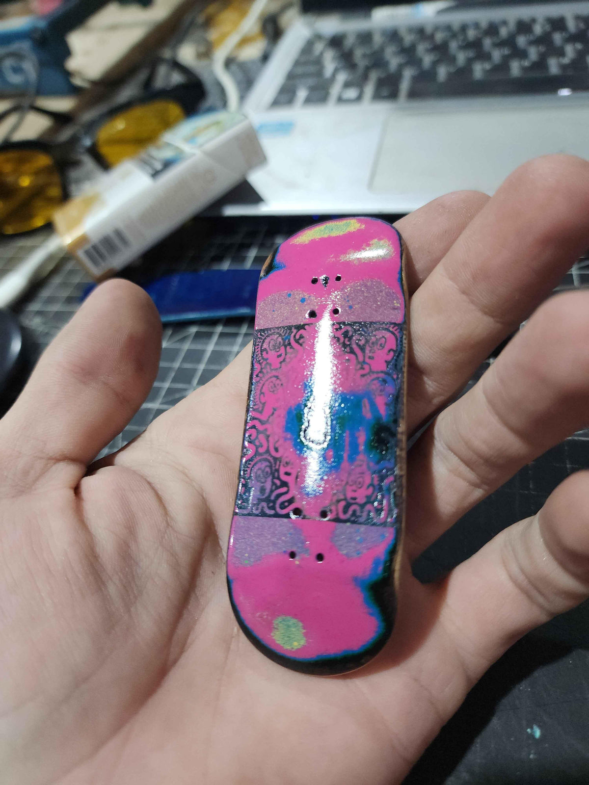 A hand holding a handmade custom fingerboard deck with a colorful pink and blue abstract design.