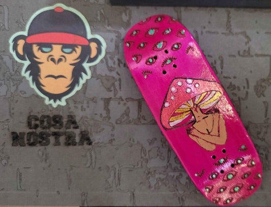Pink custom fingerboard deck with mushroom and eye graphics next to a monkey sticker.