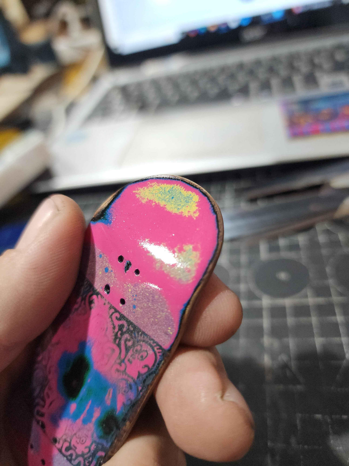 Hand holding a custom handmade pink fingerboard deck with colorful graphic design.
