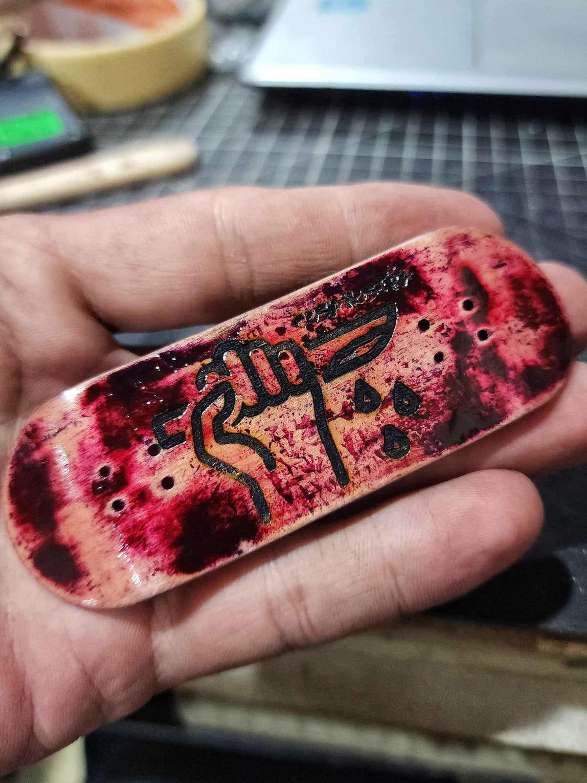 Hand holding a custom fingerboard deck with red and black abstract art and calligraphy.