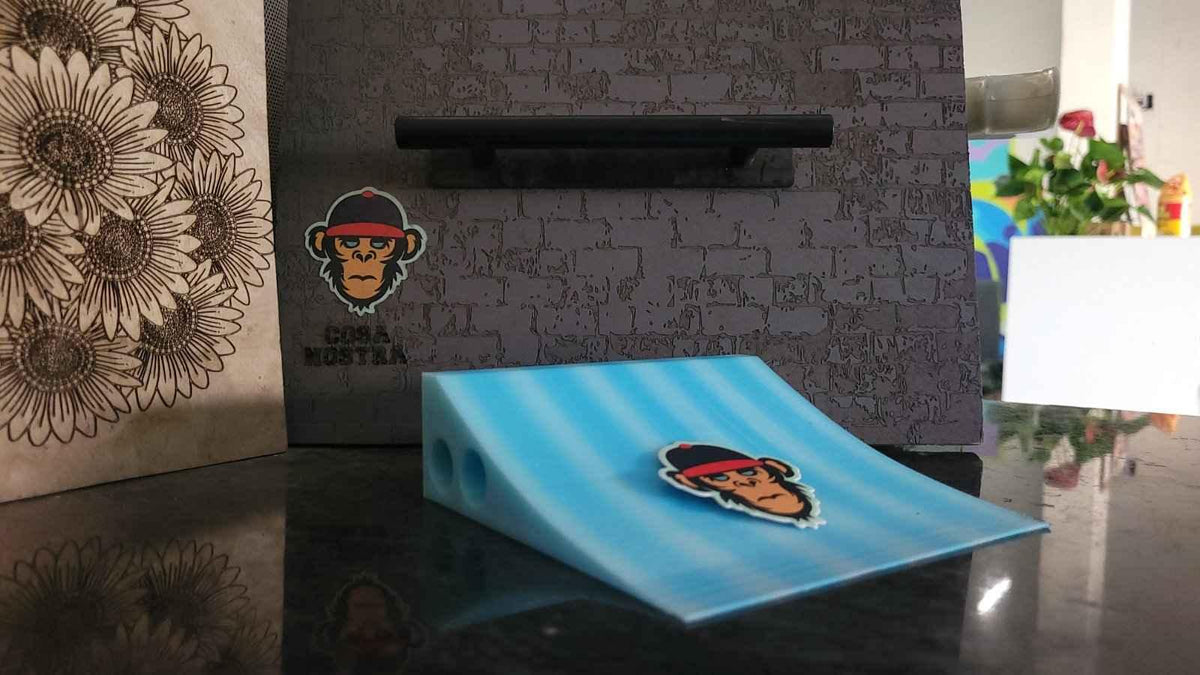 Blue fingerboard mini ramp ramp with a monkey sticker on a dark surface.