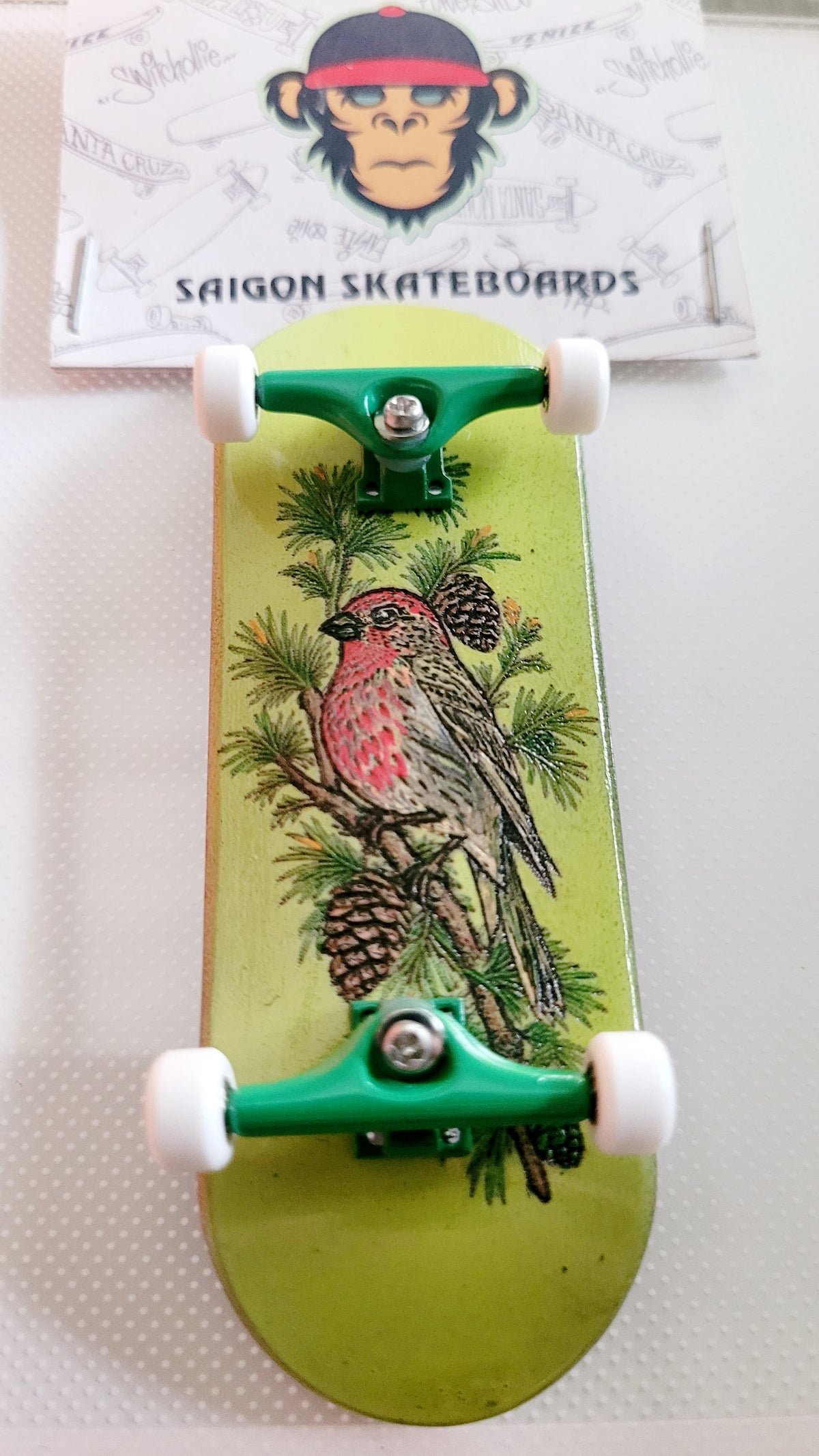 Fingerboard deck with pine-and-bird graphic, green trucks, and white wheel set