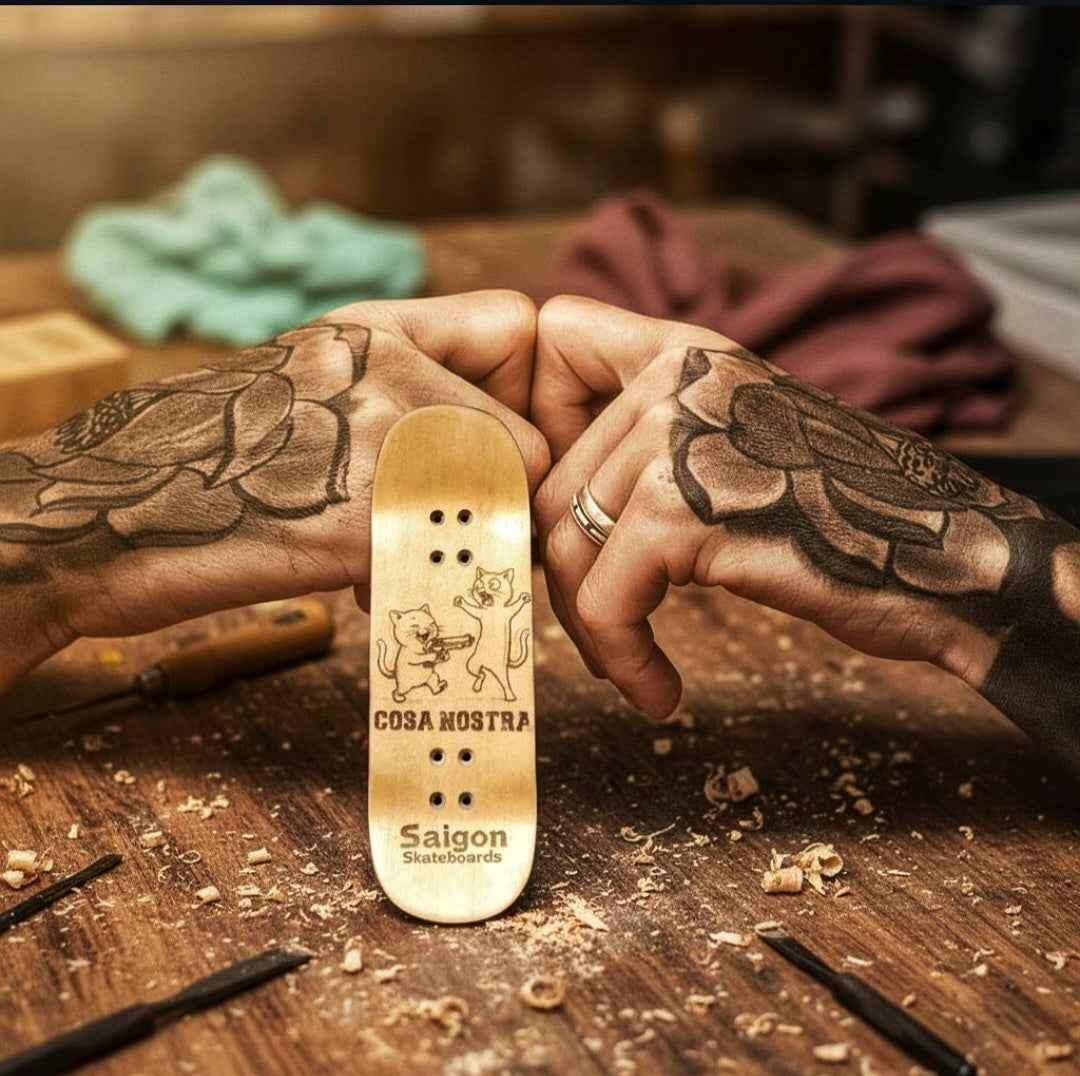 Tattooed hands holding a custom Cosa Nostra fingerboard deck on a wooden workbench.