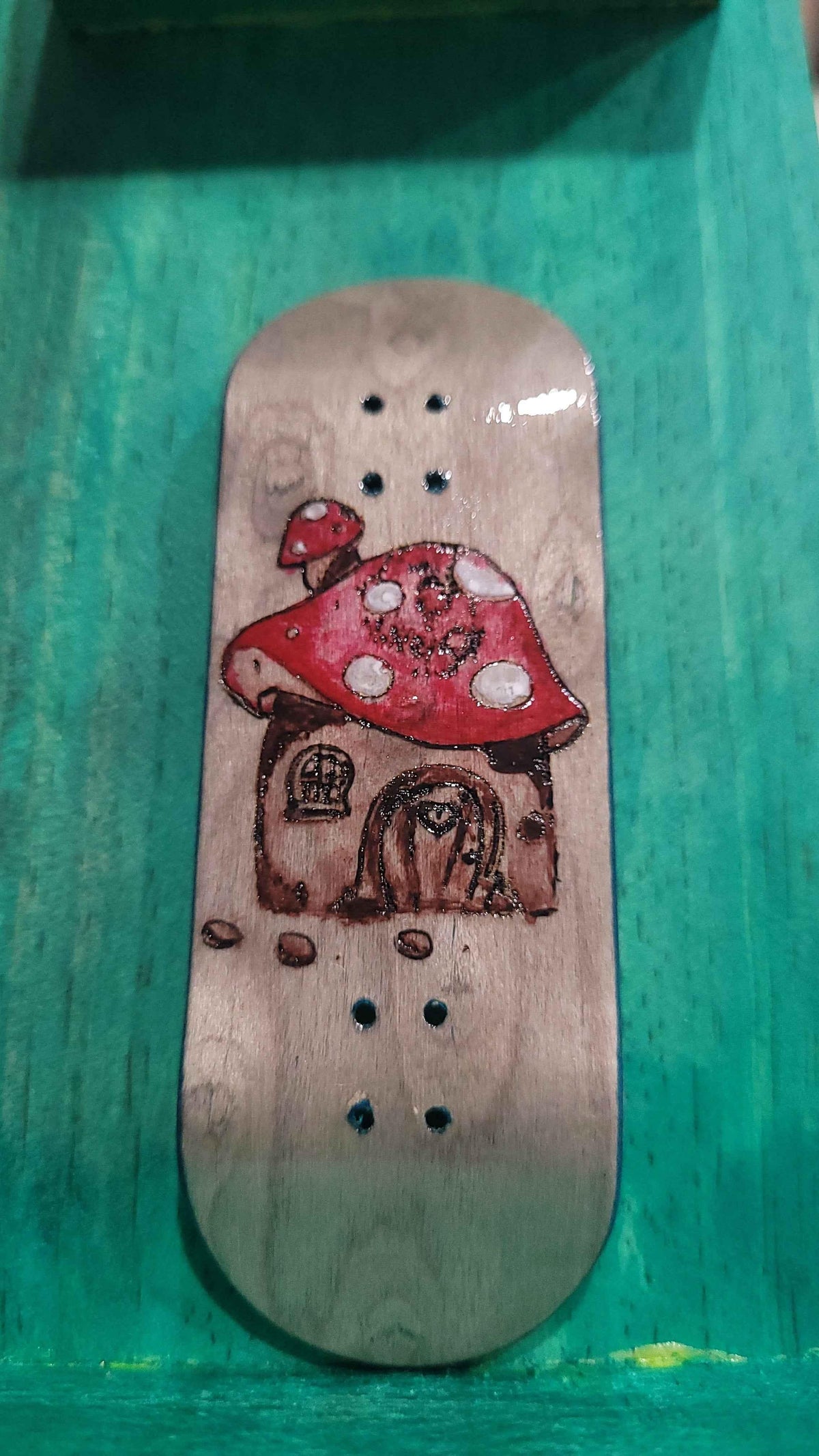 Handmade custom fingerboard deck with a red mushroom house graphic on light wood.