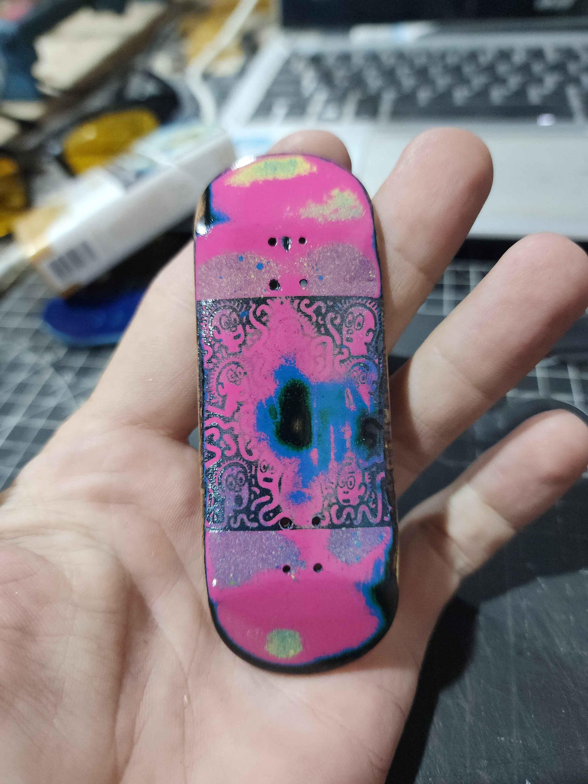 Hand holding a custom handmade fingerboard deck with pink and blue psychedelic graphics.