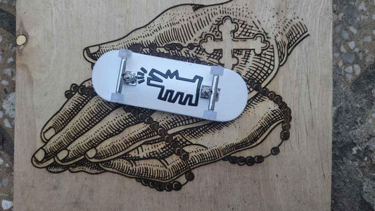 White custom fingerboard deck with black graphic resting on a wood engraving of hands and a cross.