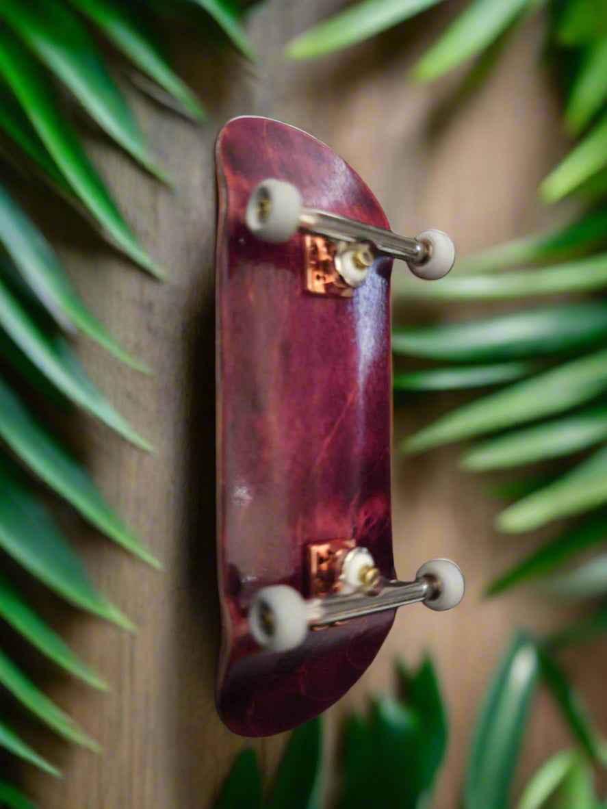 Handmade custom purple fingerboard deck with white wheels on a wooden surface with green leaves.