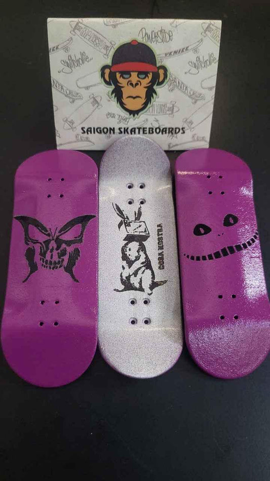 Three custom handmade fingerboard decks with graphic designs and a brand box.