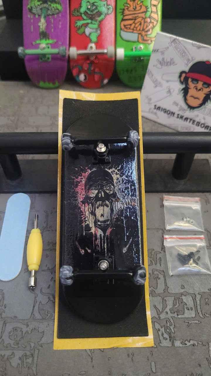 Custom black fingerboard deck with skull graphic and miniature trucks being assembled.