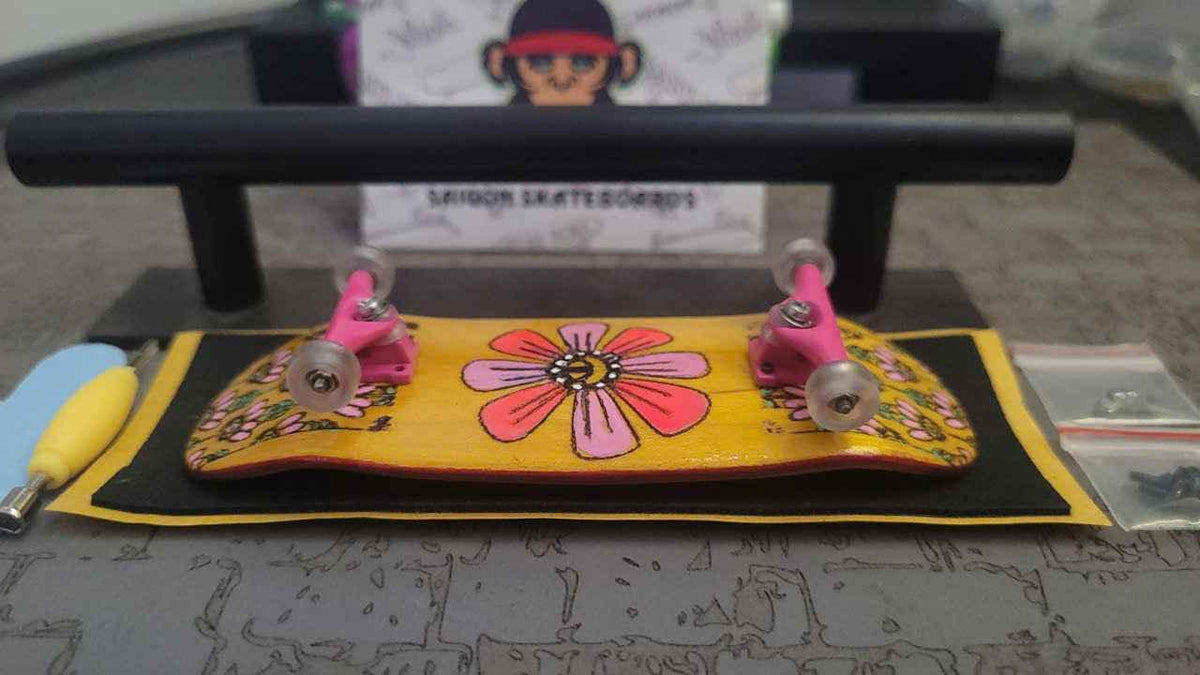 Custom handmade fingerboard with pink trucks and a yellow floral deck on a black rail.