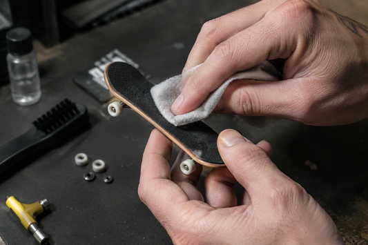Hands wiping a wooden fingerboard’s black grip surface with a cloth at a workbench.