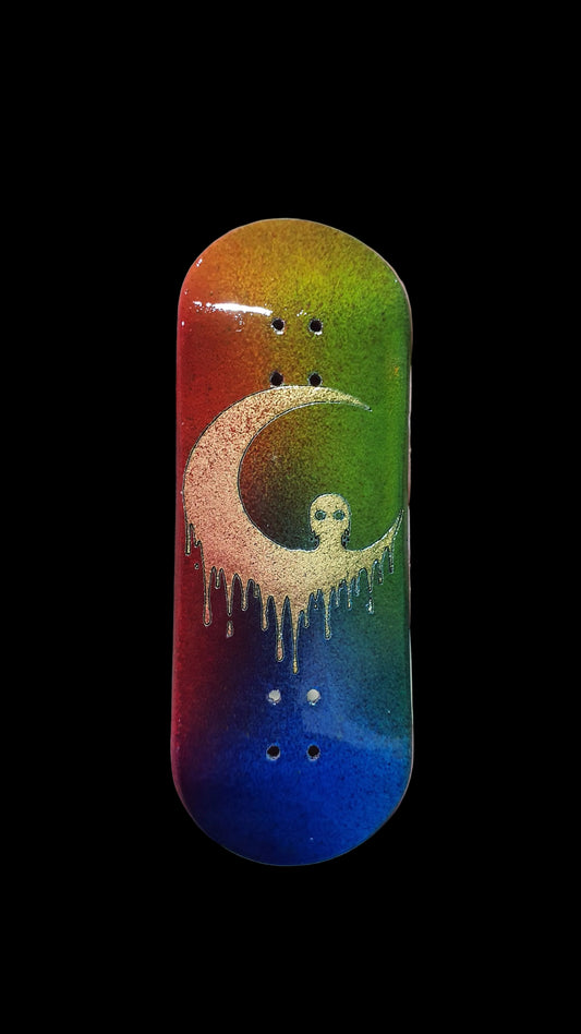 A skateboard deck with a vibrant rainbow gradient background and a central cream-colored dripping crescent moon graphic containing a small skull.