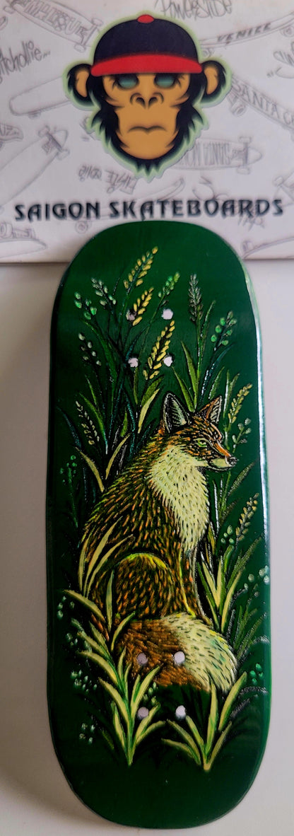 A dark green Fingerboard deck featuring a hand-painted illustration of a fox in tall grass, with a Saigon Skateboards logo card visible behind it.