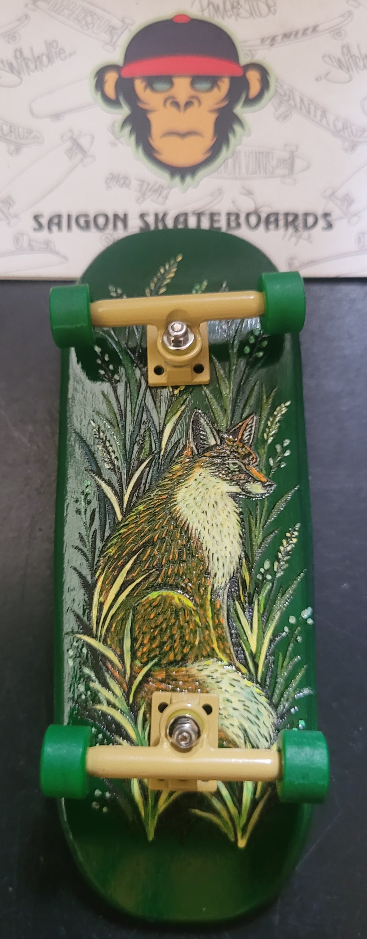 A green fingerboard deck featuring a detailed illustration of a fox sitting in tall grass, with green wheels and beige trucks, displayed on a dark surface with a Saigon Skateboards logo card in the background.