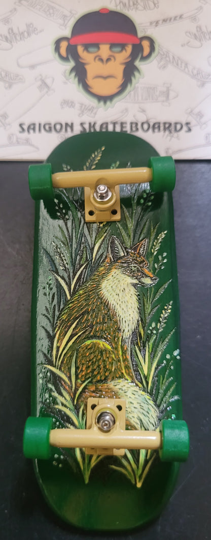 A green fingerboard deck featuring a detailed illustration of a fox sitting in tall grass, with green wheels and beige trucks, displayed on a dark surface with a Saigon Skateboards logo card in the background.