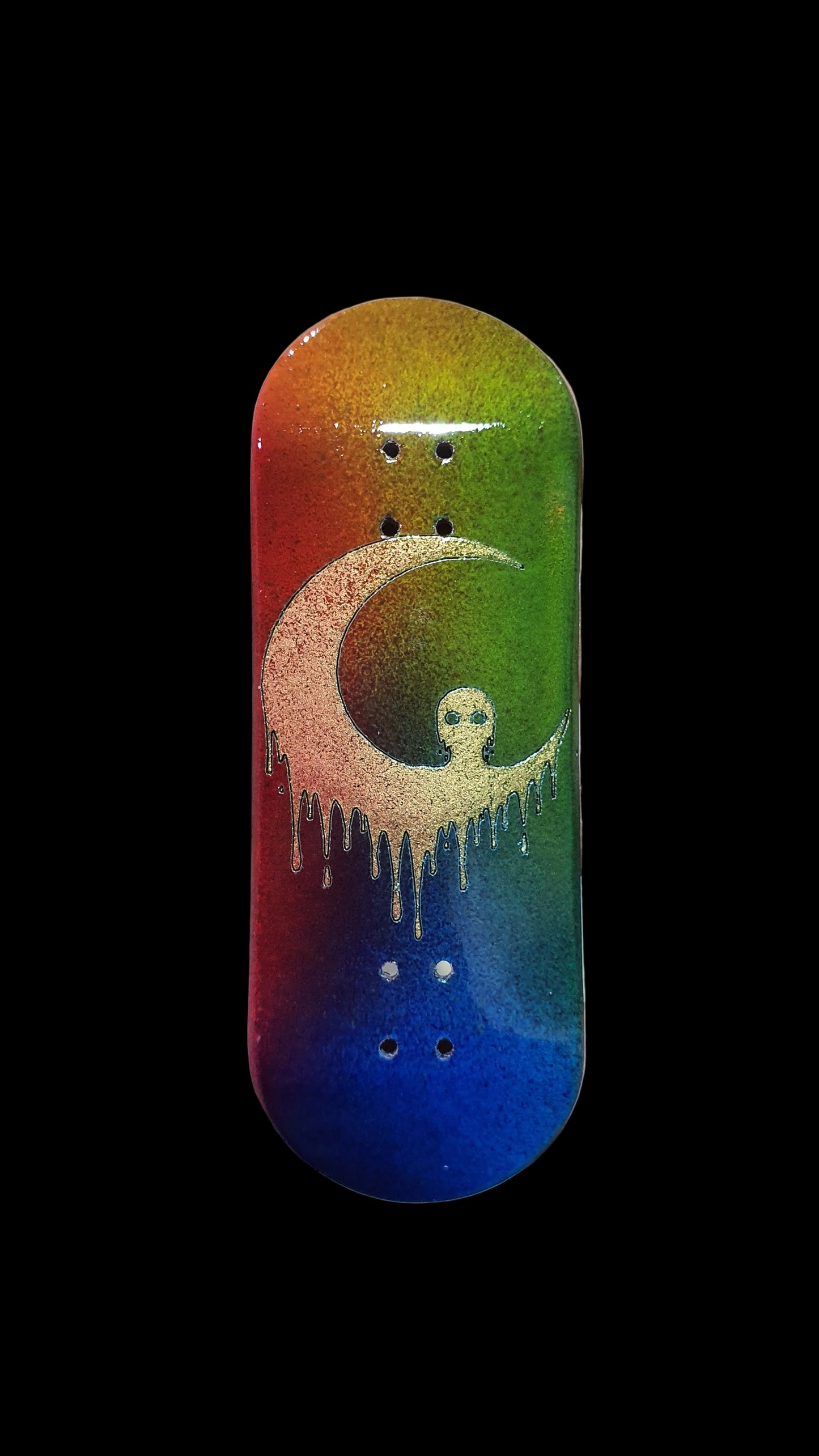 A hand holding a miniature skateboard deck featuring a vibrant rainbow gradient background with a gold glitter crescent moon and ghost design.
