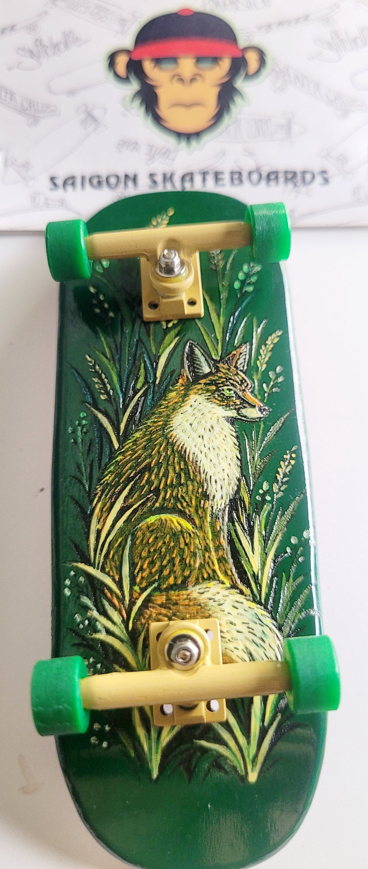 A miniature fingerboard with a green deck featuring an illustrated fox in tall grass, green wheels, and light wood trucks. A Saigon Skateboards logo card is visible in the background.