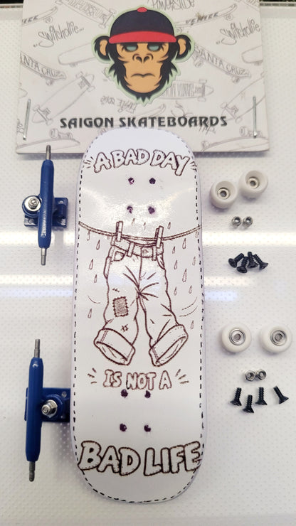A complete skateboard deck set laid out on a white surface, featuring a white skateboard deck with a brown line art illustration of pants and the text 'ABADAY IS NOT A BAD LIFE', blue trucks, white wheels, and screws. A branded 'SAIGON SKATEBOARDS' card with a monkey graphic is visible in the background.
