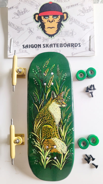 A complete fingerboard deck set from Saigon Skateboards, featuring a green deck with a detailed illustration of a fox in a field, accompanied by two yellow trucks, two green wheels, and black hardware, arranged on a white surface with the brand's logo card in the background.