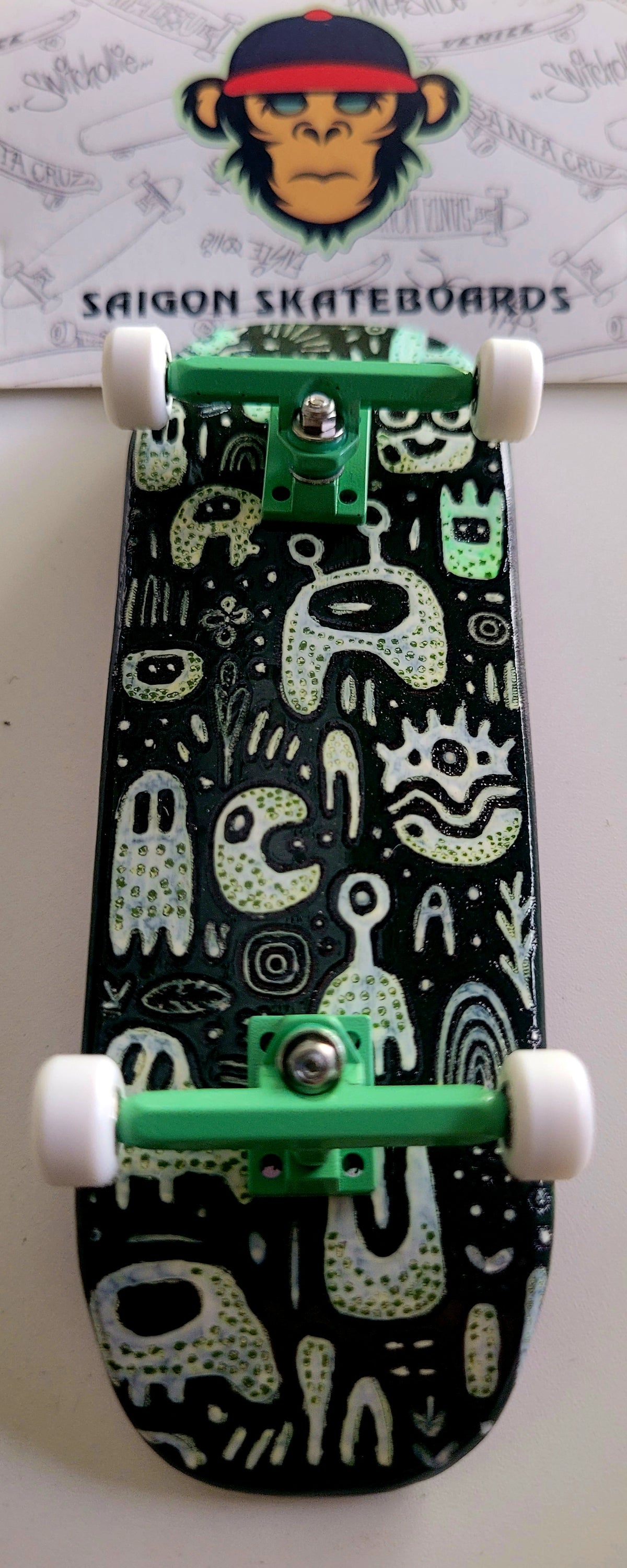 Black medium fingerboard with white and green monster doodle art, green trucks, and white wheels.
