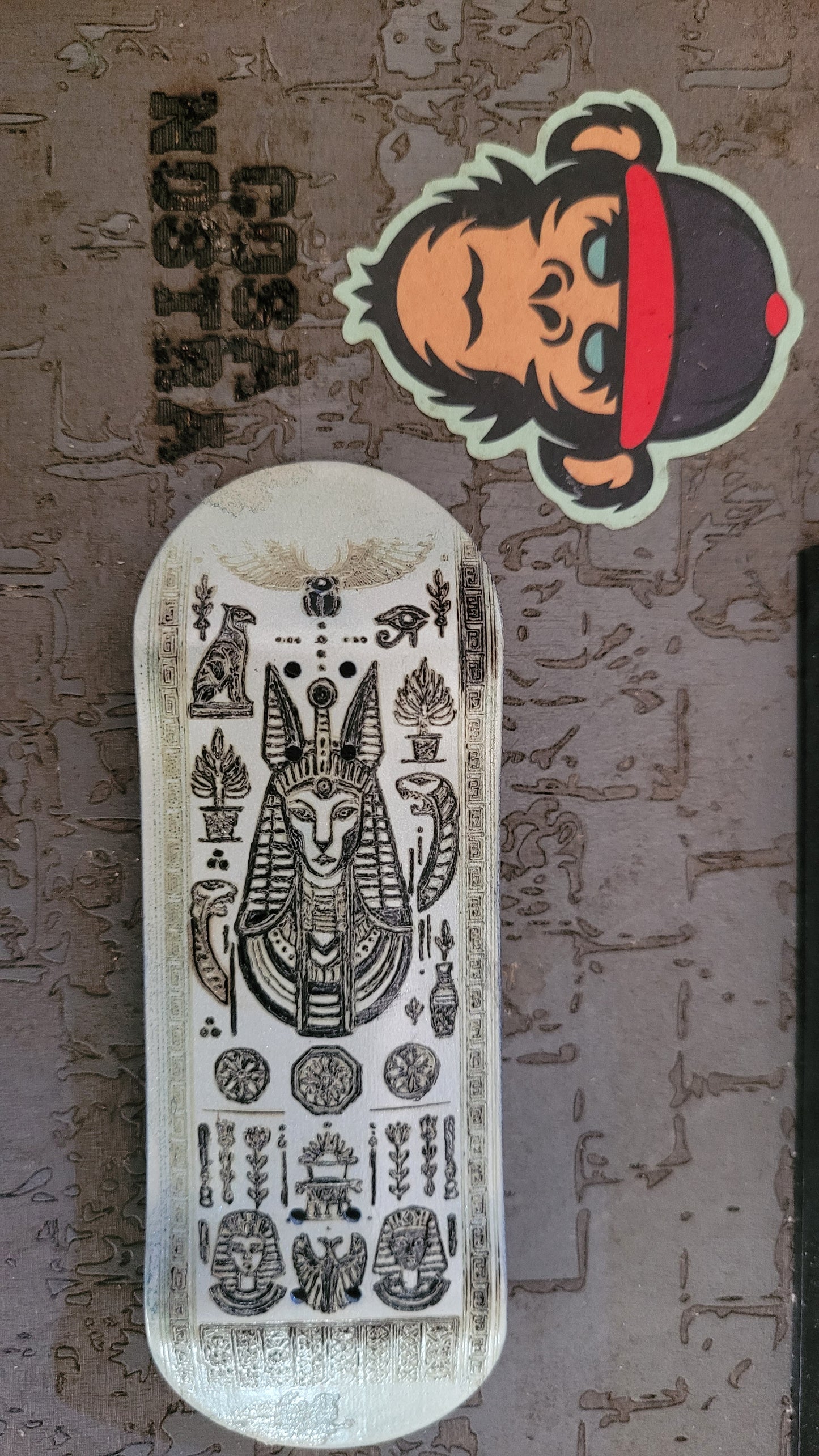 Sticker of a monkey with a cap and 'Cosa Nostra' text next to a skateboard deck with intricate designs on a textured surface.