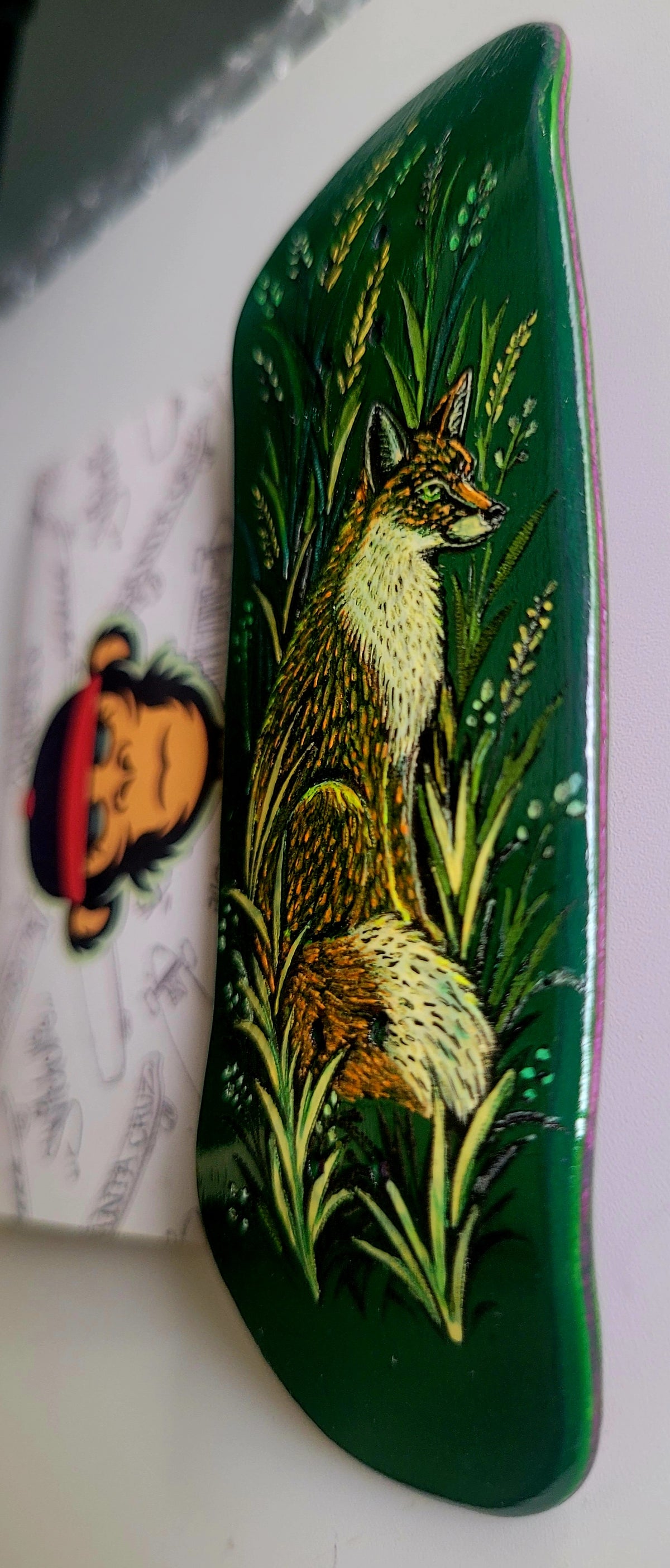 A dark green fingerboard deck featuring a hand-painted illustration of a fox sitting among tall green and yellow grass. A white sticker with a cartoon monkey is partially visible on the left against a light background.