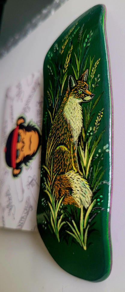 A dark green fingerboard deck featuring a hand-painted illustration of a fox sitting among tall green and yellow grass. A white sticker with a cartoon monkey is partially visible on the left against a light background.