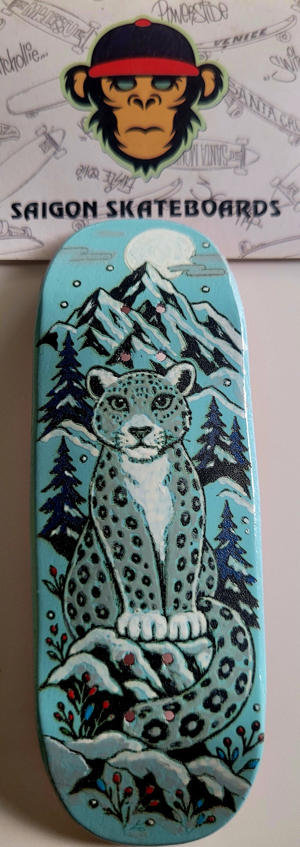 Teal finger skateboard deck with snow leopard and pine trees graphic
