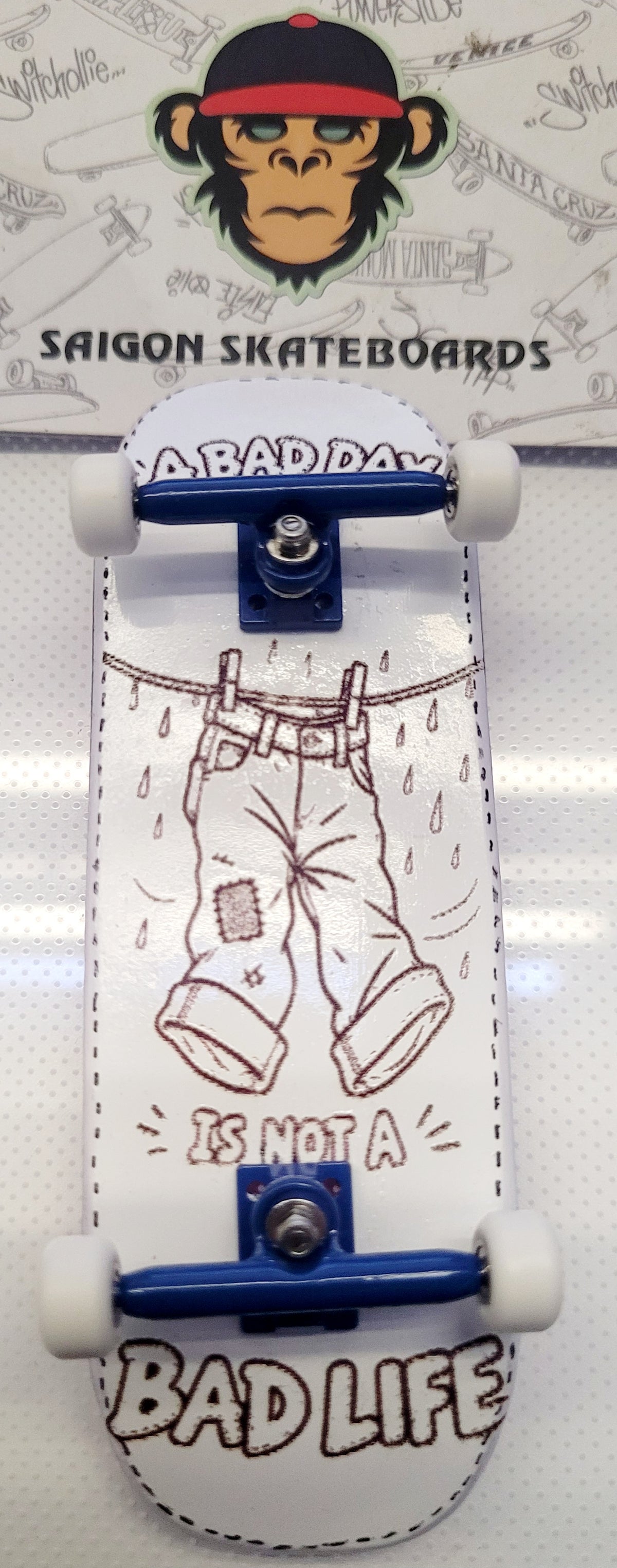 A miniature skateboard with a white deck featuring a brown line art illustration of pants on a clothesline with the text 'IS NOT A BAD DAY' and 'BAD LIFE'. The skateboard has blue metal trucks and white wheels, photographed against a white background with a branded 'SAIGON SKATEBOARDS' card behind it.