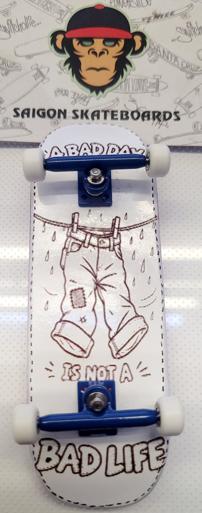A miniature skateboard with a white deck featuring a brown line art illustration of pants on a clothesline with the text 'IS NOT A BAD DAY' and 'BAD LIFE'. The skateboard has blue metal trucks and white wheels, photographed against a white background with a branded 'SAIGON SKATEBOARDS' card behind it.