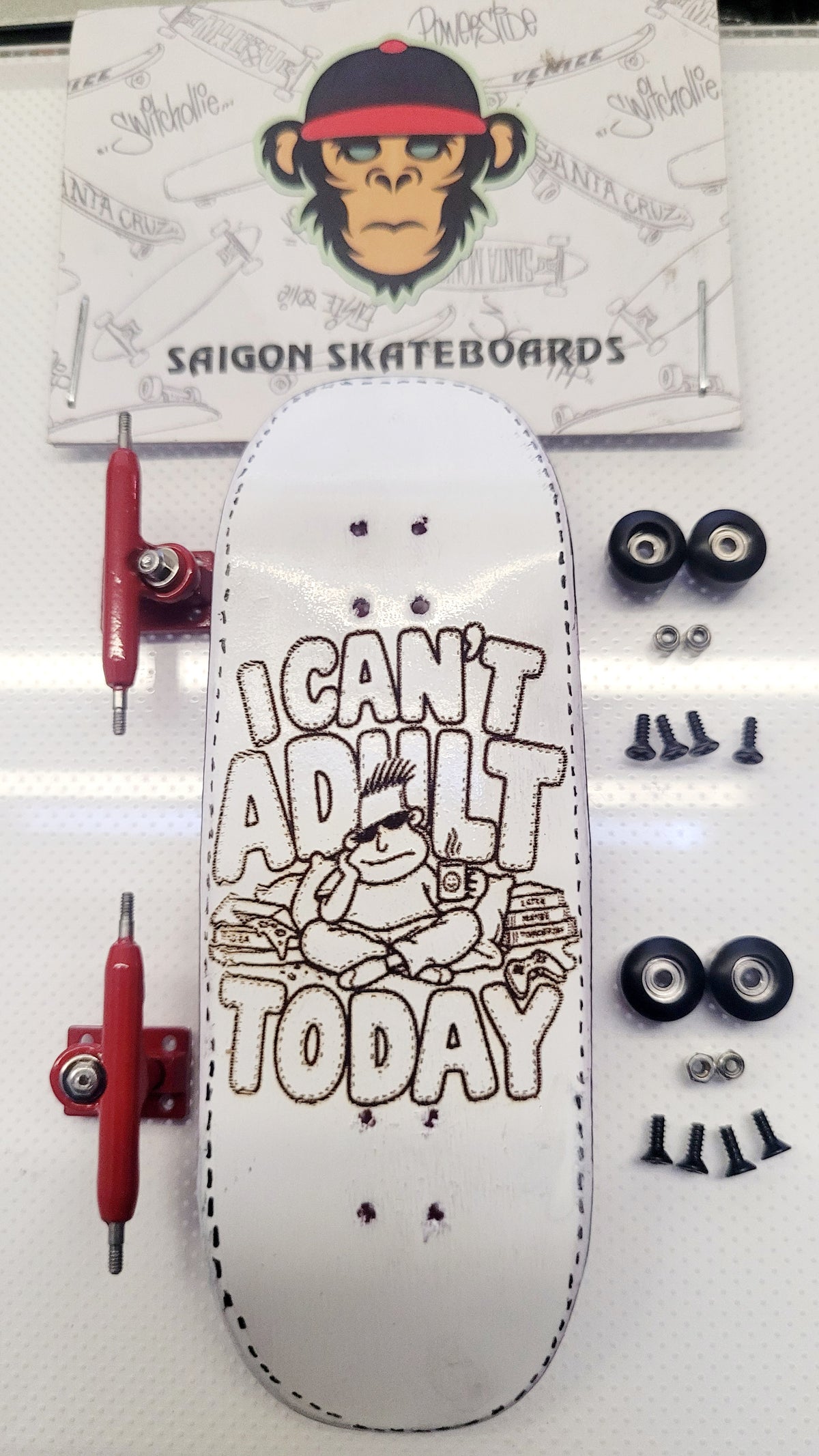 A white skateboard deck with a brown graphic reading 'I CAN'T ADULT TODAY' and a monkey illustration, displayed with red trucks, black wheels, and mounting hardware on a white surface. A branded Saigon Skateboards card is visible in the background.