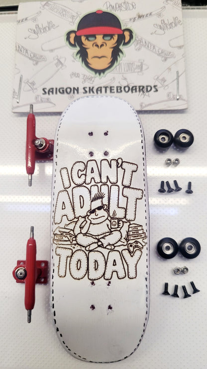 A white skateboard deck with a brown graphic reading 'I CAN'T ADULT TODAY' and a monkey illustration, displayed with red trucks, black wheels, and mounting hardware on a white surface. A branded Saigon Skateboards card is visible in the background.