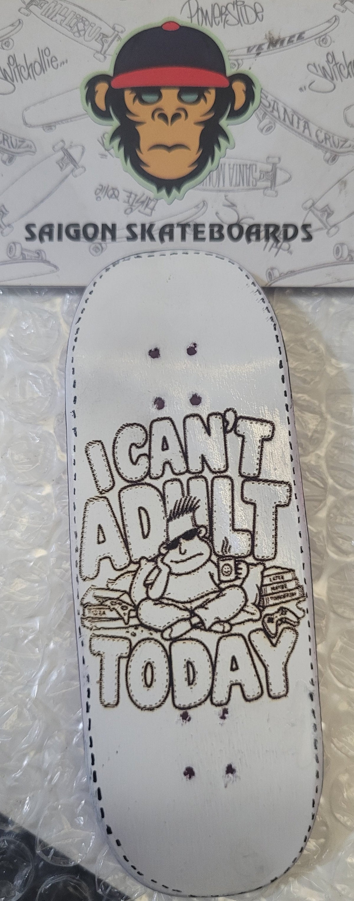A miniature white skateboard deck with a hand-drawn style graphic of a cartoon character and the text 'I CAN'T ADULT TODAY' in black. The deck is displayed on bubble wrap next to its branded cardboard backing card featuring a monkey illustration and the brand name 'SAIGON SKATEBOARDS'.