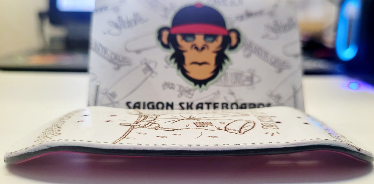 A white leather wallet with brown line art illustrations and 'SAIGON SKATEBOARDS' text, shown in front of its branded white pouch packaging featuring a monkey graphic.