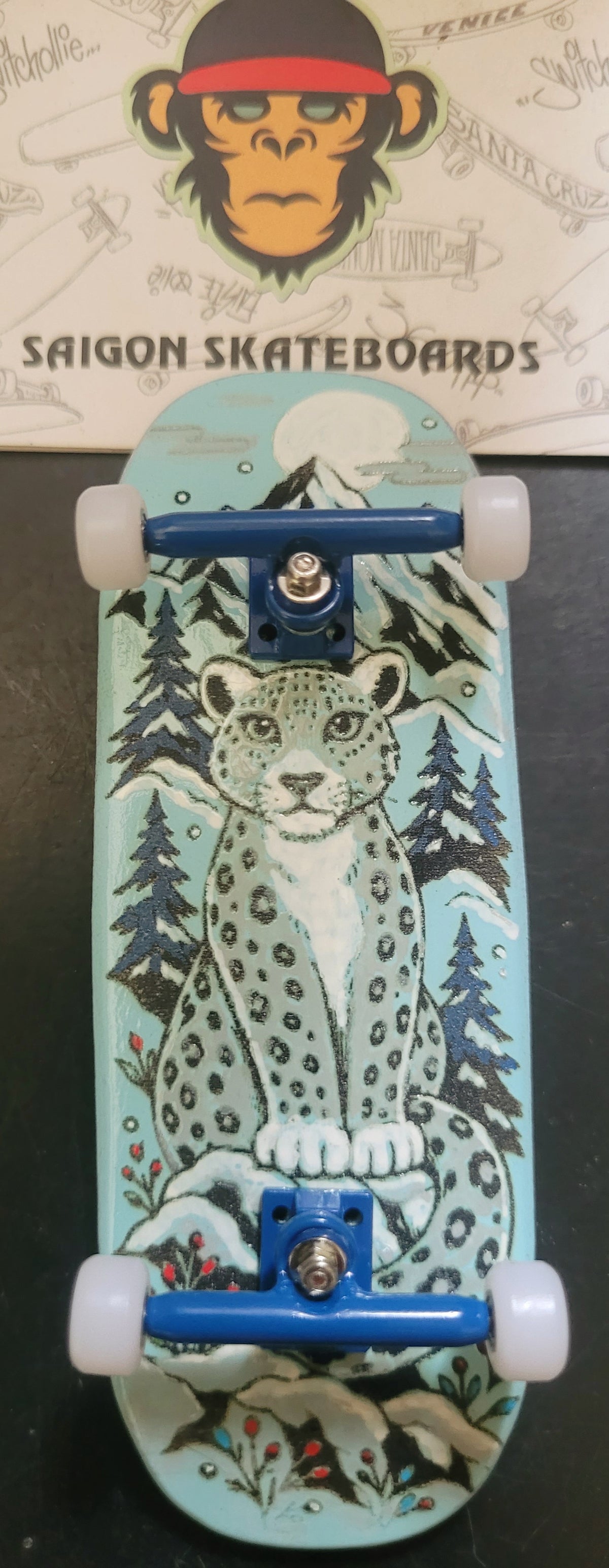 Teal finger skateboard with snow leopard graphic, blue trucks and four white wheels