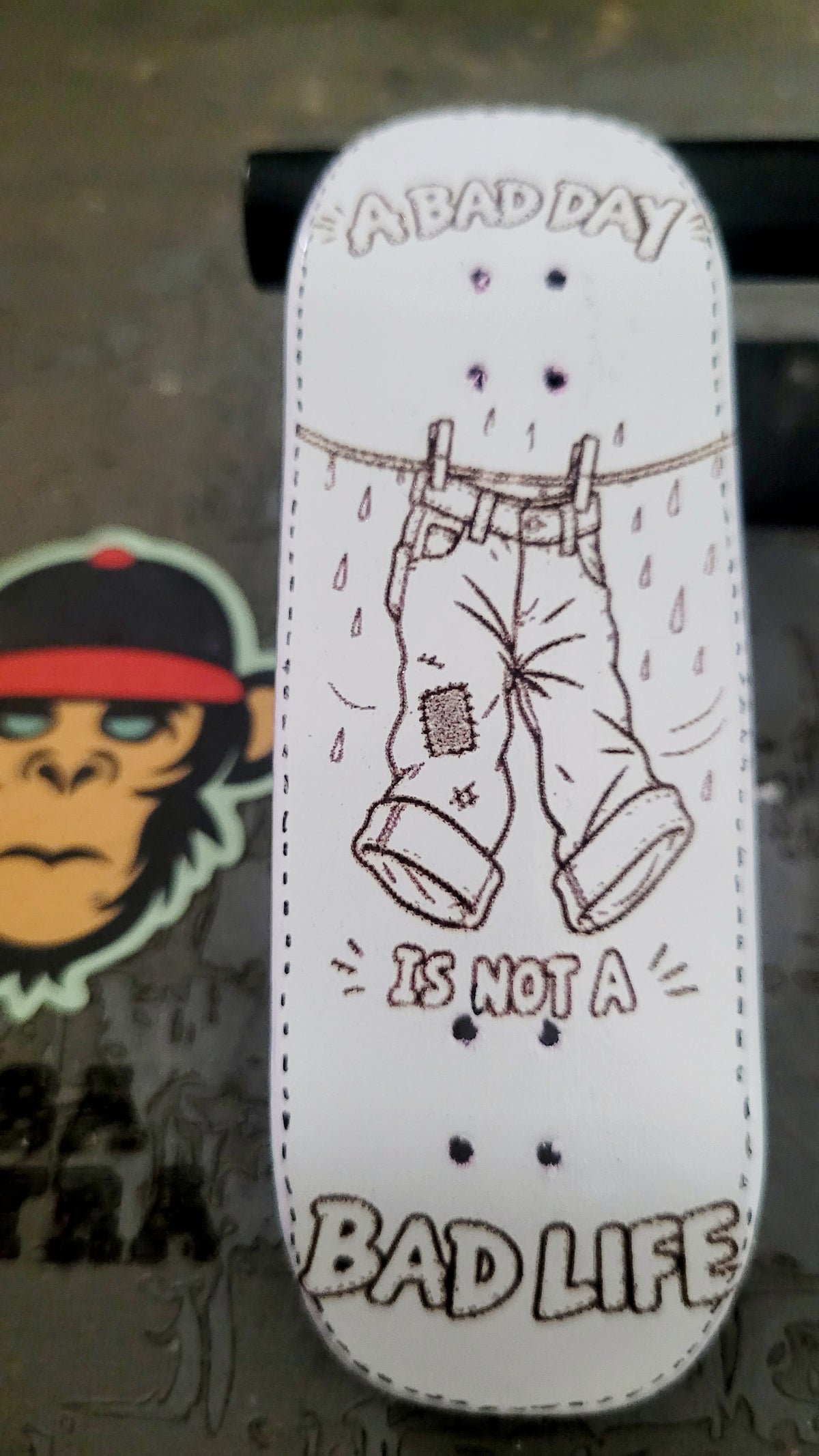 A white skateboard deck with a hand-drawn illustration of a pair of pants hanging on a clothesline with raindrops, surrounded by the text 'A BAD DAY IS NOT A BAD LIFE'. A monkey sticker is partially visible on the left on a dark textured surface.