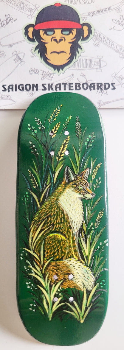 A green Fingerboard deck featuring a painted illustration of a fox sitting among tall grass, shown against a white background with a branded sticker above it.